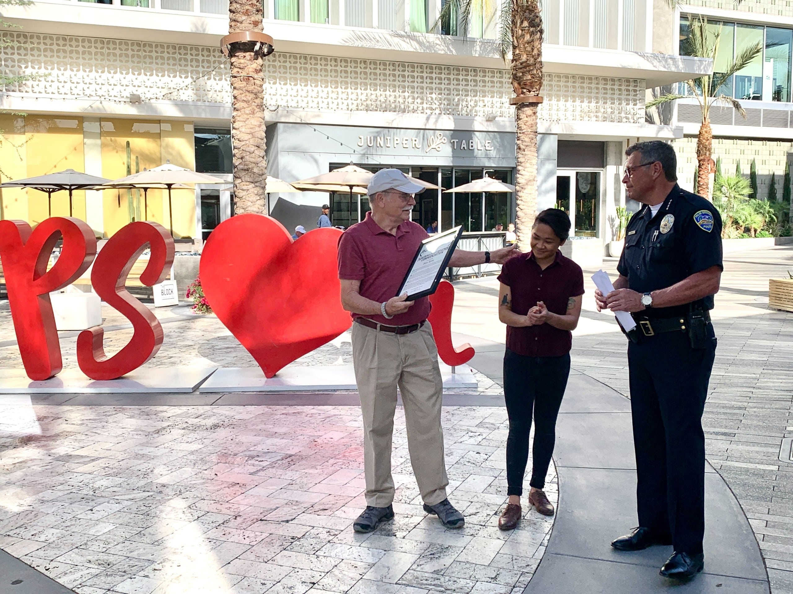 A man holds a plaque and smiles next to a woman and a police chief in front of a large red sign that says "PS heart you"
