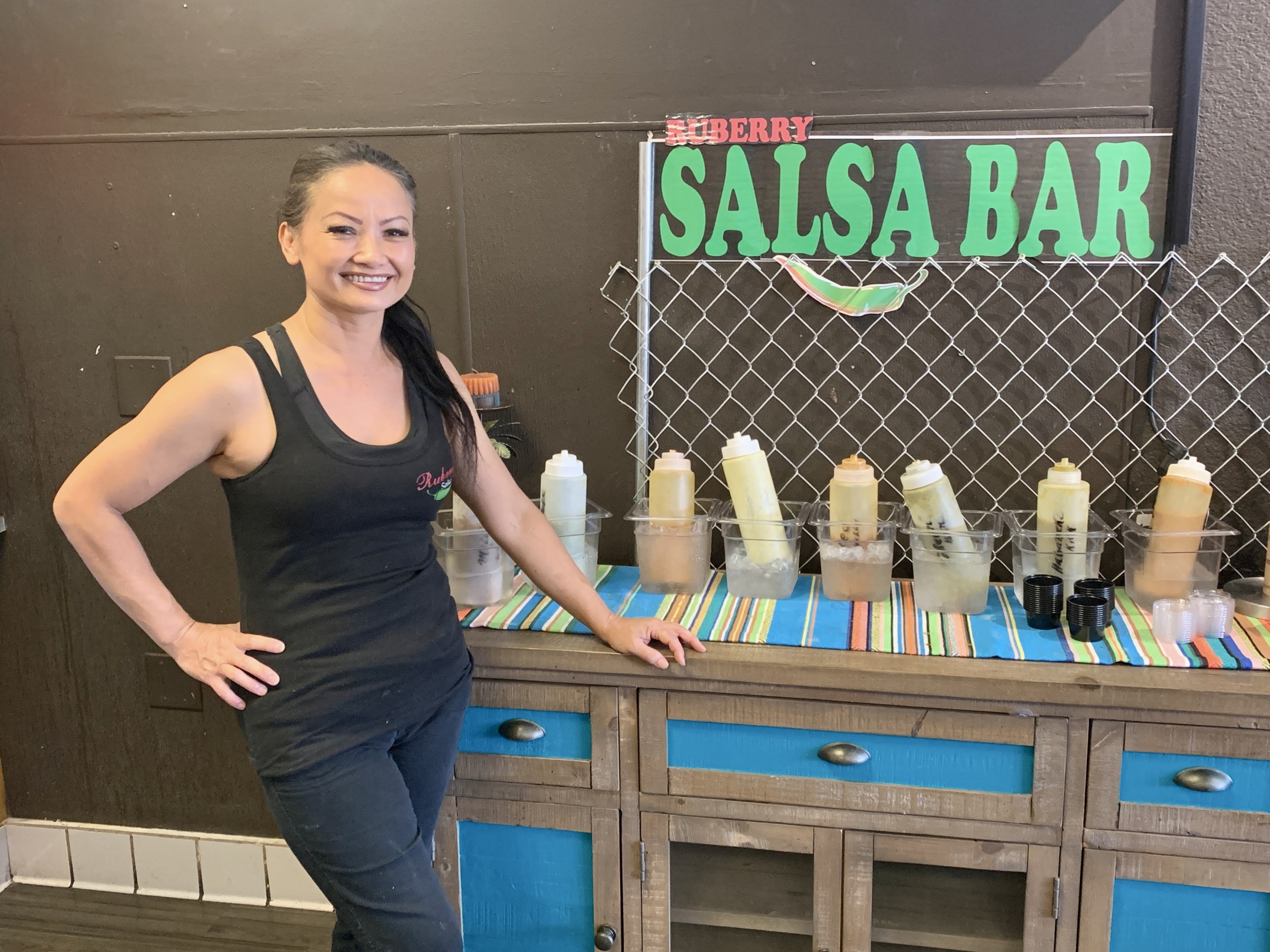 A woman wearing black smiles and poses in front of a sign that says Salsa Bar and a table with salsa bottles